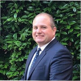 Man in suit smiling against green leafy background.